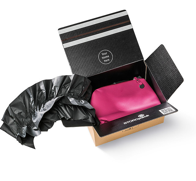 Ein Karton ist innen schwarz gefärbt und beinhaltet eine pinke Tasche, die von schwarzem Papierpolster umgeben ist.